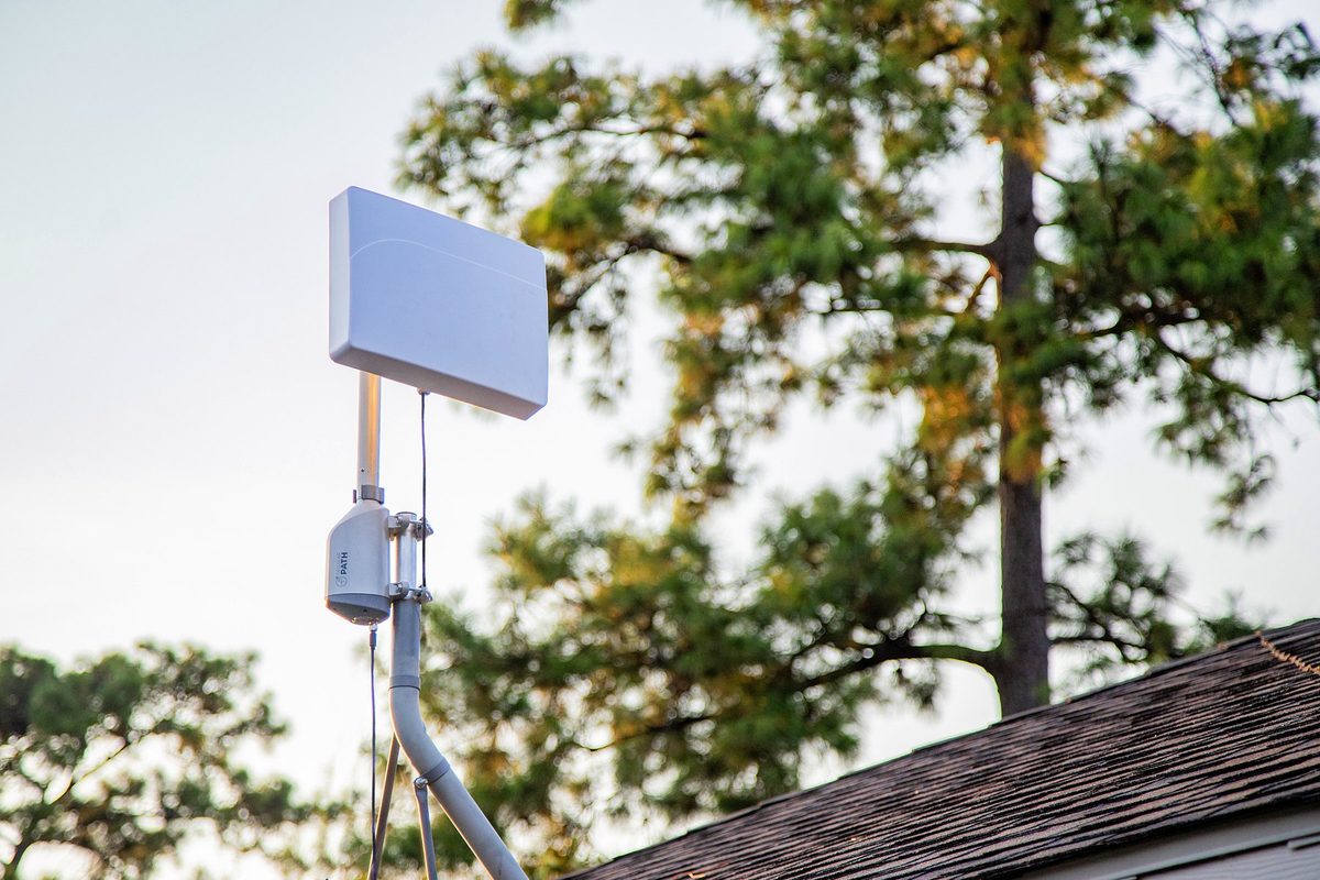 A SOCA rotator on a roof mast carrying a flat panel antenna, with trees and a roofline behind it.
