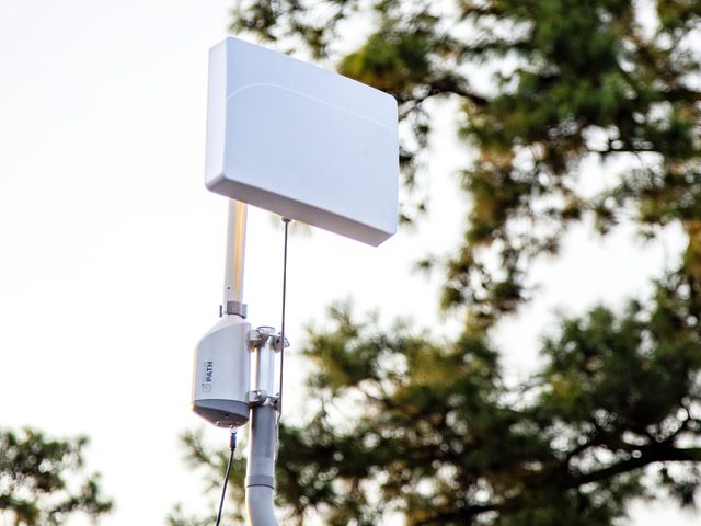 A SOCA rotator on a mast carrying a flat panel antenna, photographed from below against trees and sky.